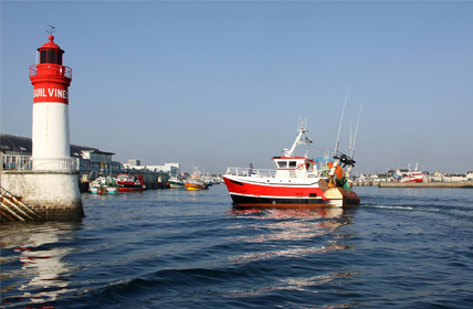 Photo d'un bateau de pêche entrant au port devant le phare, rouge et blanc tous deux